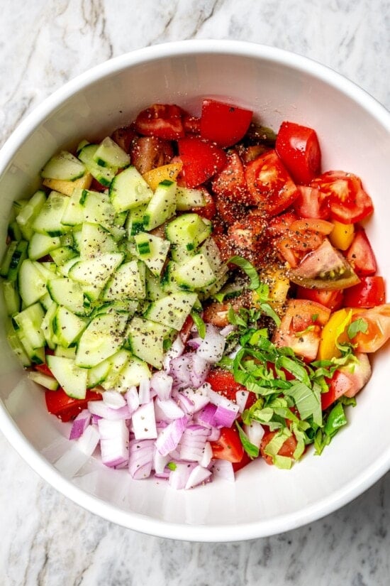 Overhead view of basil, tomatoes, red onions, and cucumbers in bowl for panzanella