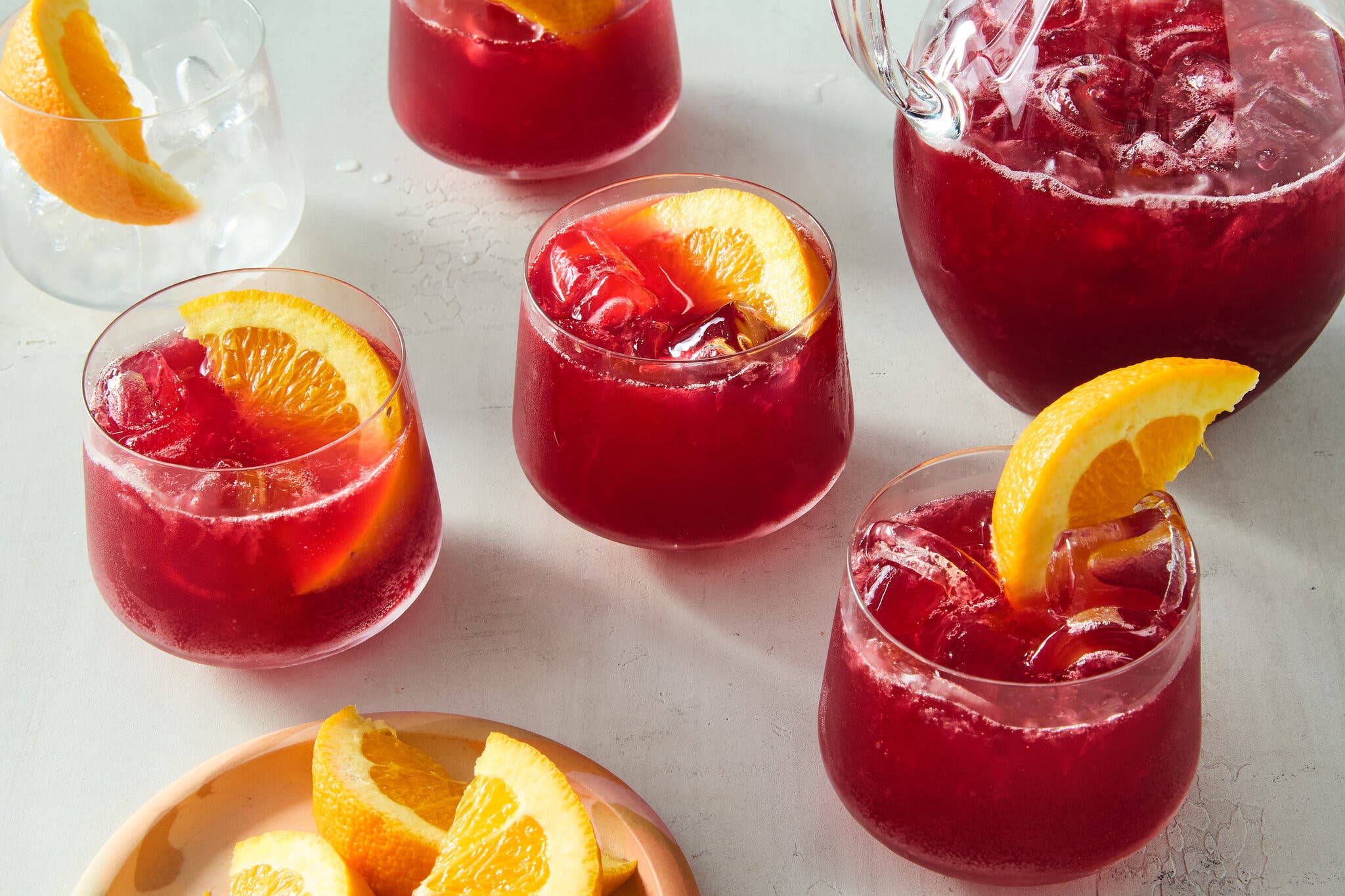 Short glasses and a pitcher are filled with red Negroni, ice and orange wedges.