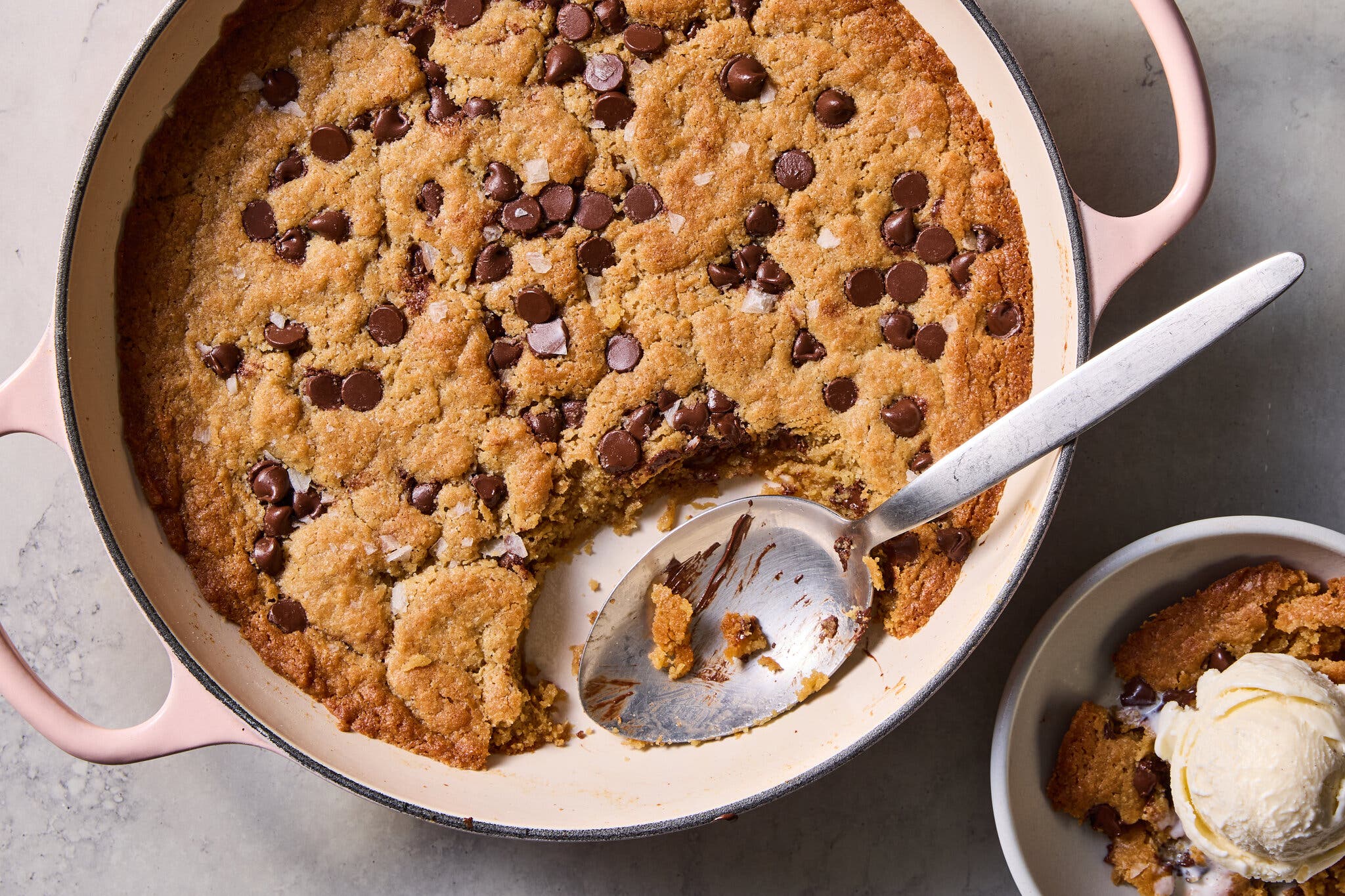 A chocolate chip skillet cookie with a scoop taken out, next to a small bowl of ice cream.
