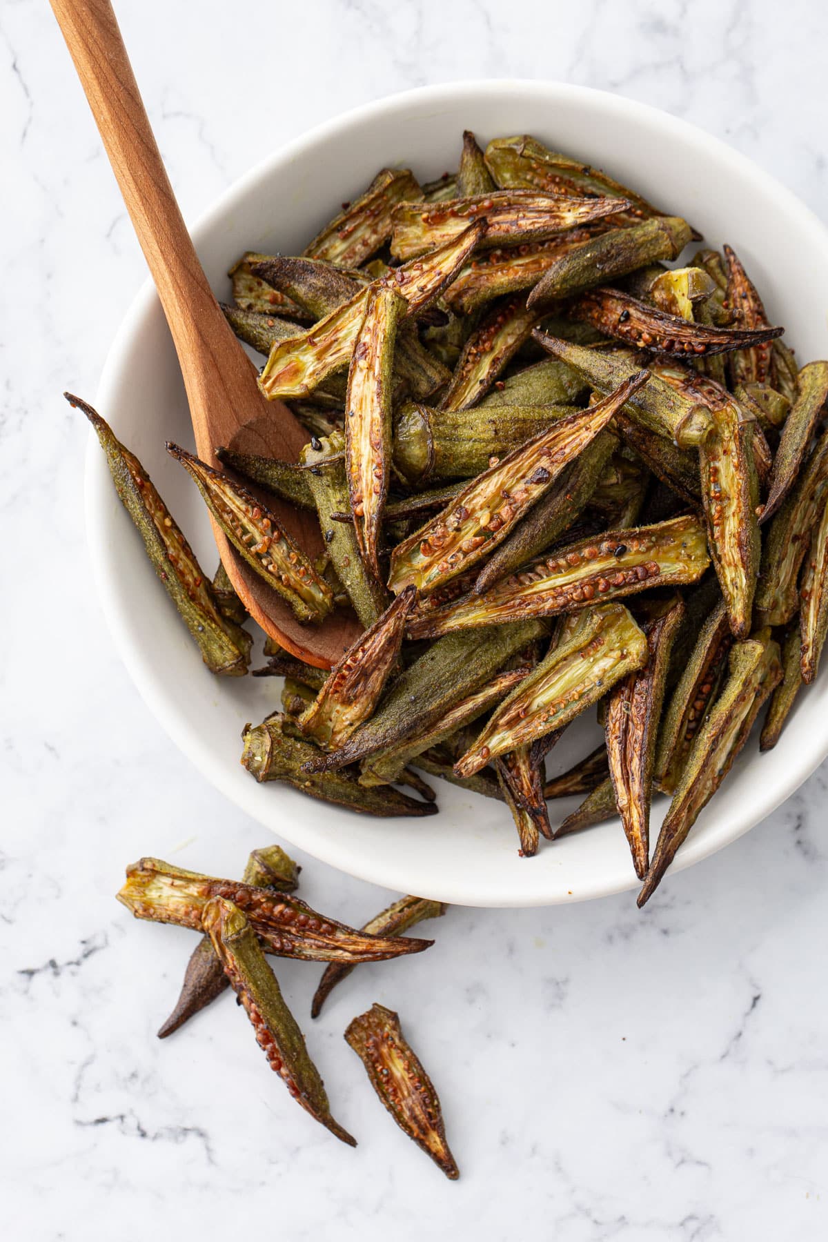 Overhead view, white bowl filled with Crispy Oven-Roasted Okra and a wooden spoon, a few okra on the marble countertop.