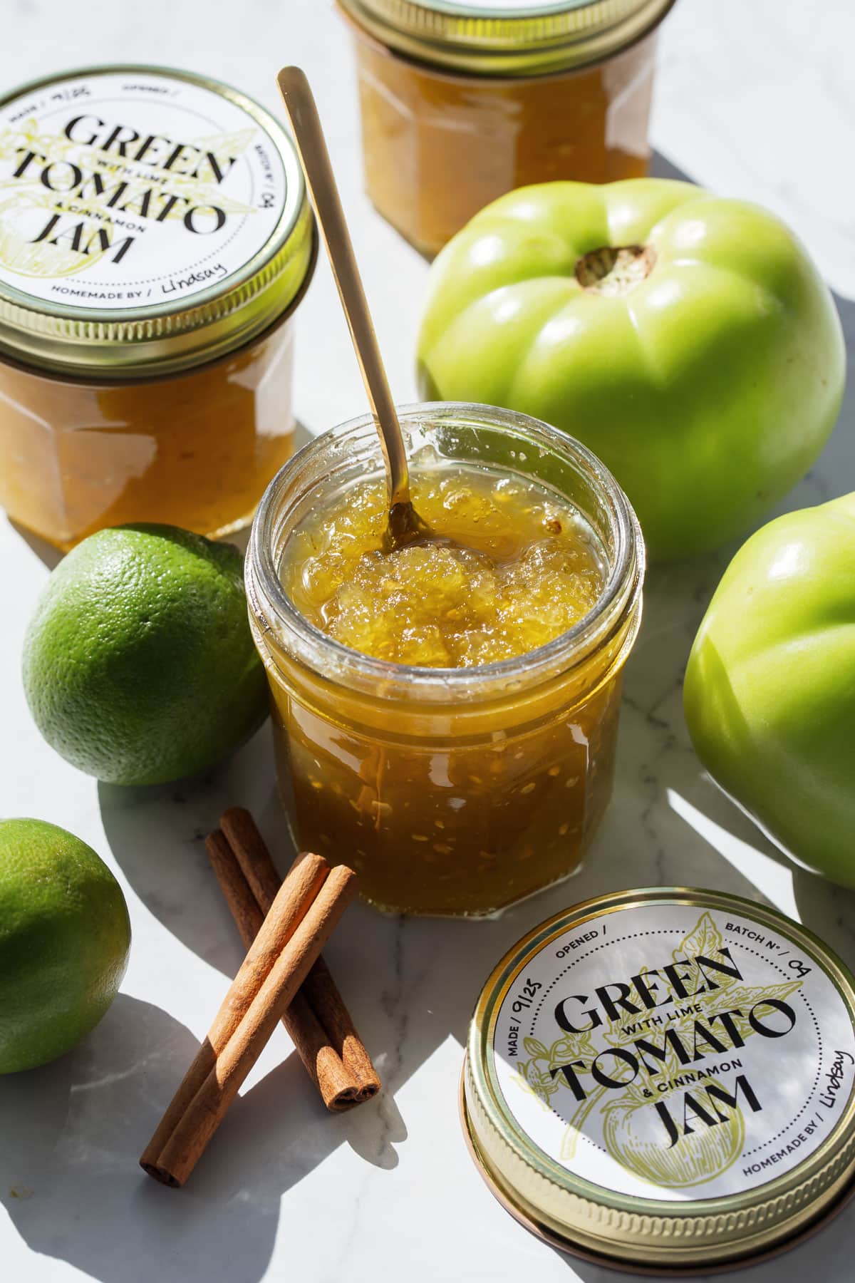 Jars of Green Tomato Jam on a marble background with whole green tomatoes, limes, and cinnamon sticks surrounding an open jar.