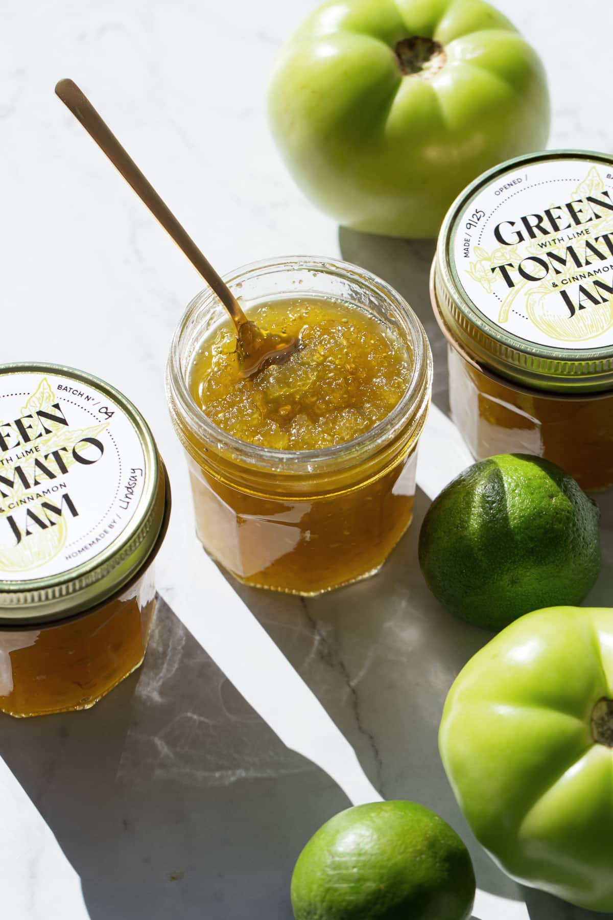 Three jars of Green Tomato Jam, one jar open with a spoon, and raw ingredients (tomatoes and limes) scattered around.