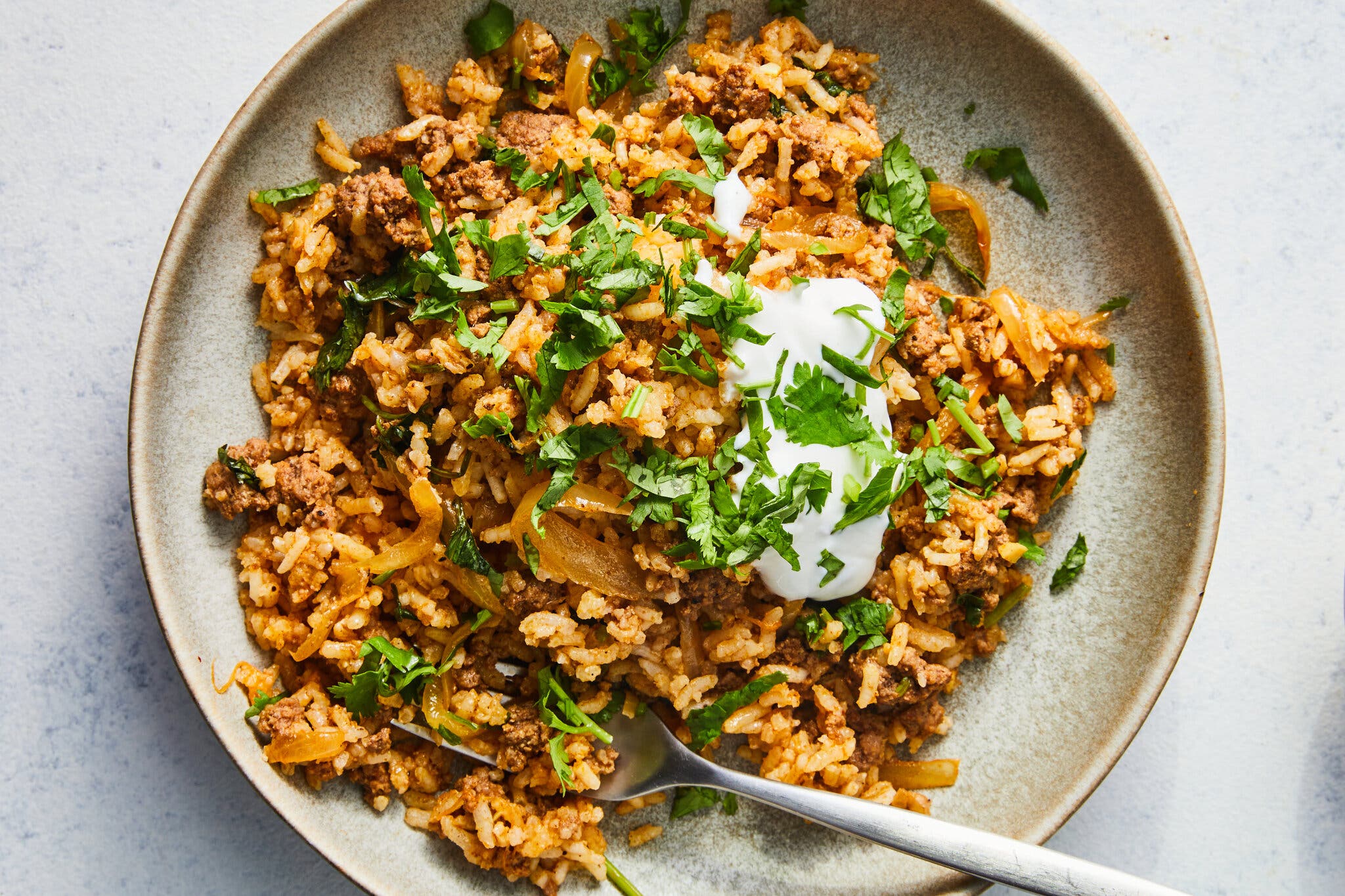 A light tan plate holds a portion of browned ground beef mixed with reddish-orange rice, topped with a pile of bright green cilantro and a small scoop of yogurt.