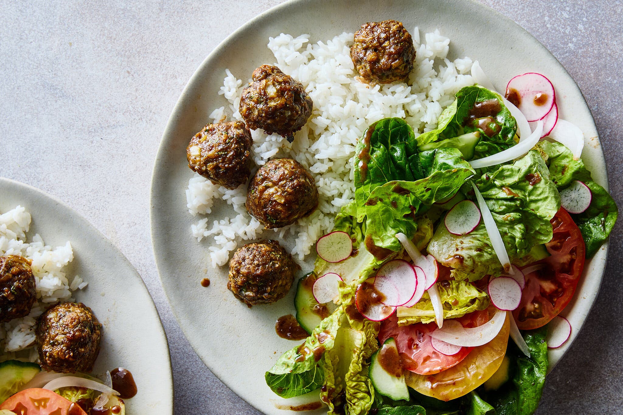 An overhead close-up of a beige plate featuring five dark, glazed meatballs and white rice on the left, and a fresh green salad with pink radishes, yellow tomatoes, and a brown dressing on the right.