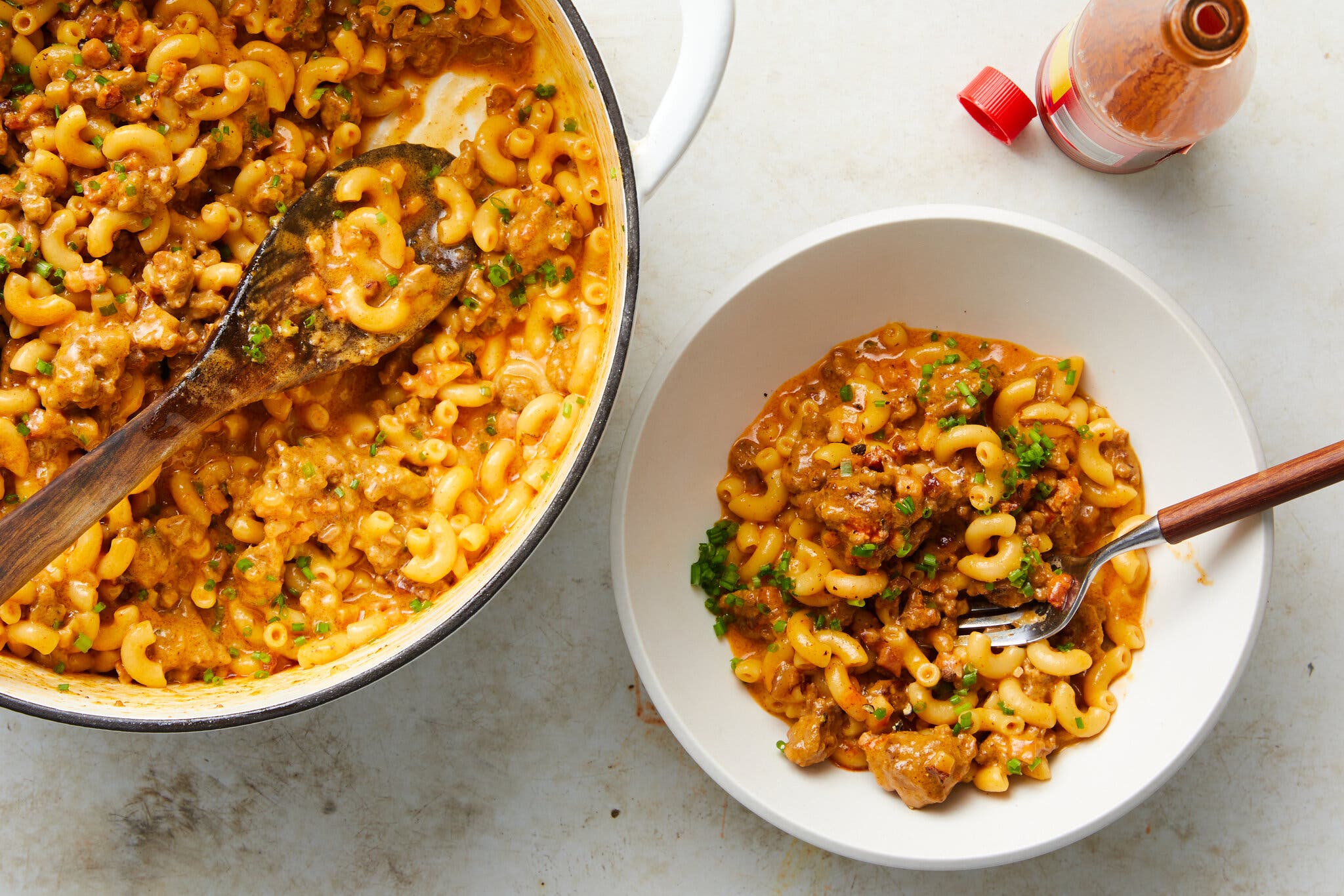 An overhead view of a large pot of cheesy beef and elbow macaroni, with a wooden spoon, alongside a single white bowl of the same dish with a fork inserted.