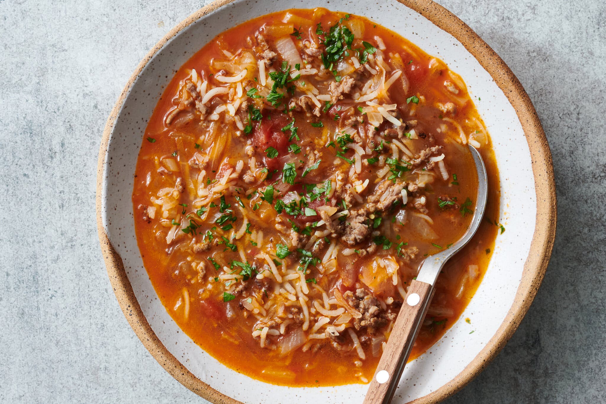 A creamy white and tan ceramic bowl holds a bright reddish-orange soup with shredded cabbage, ground beef, rice, and tomatoes, garnished with chopped green parsley; a spoon rests in the bowl.