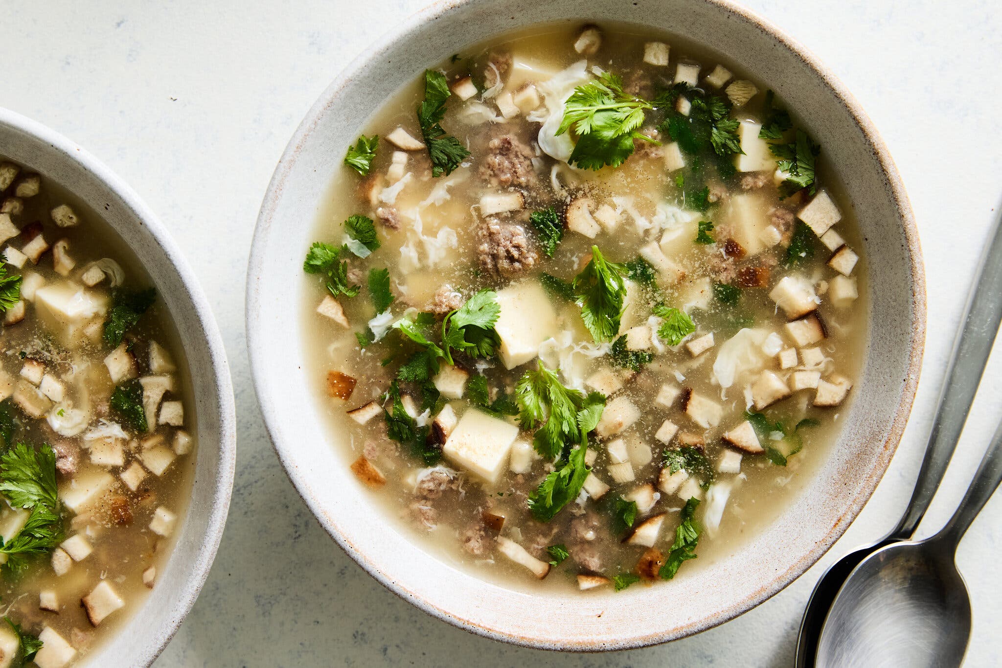 Two ceramic bowls of light brown broth, featuring ground beef, floating egg ribbons, white tofu cubes, brown mushrooms, and fresh green cilantro; two silver spoons rest beside the bowl on the right.