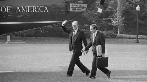 Getty Images President Gerald Ford waves as he walks past the presidential helicopter with Dick Cheney walking next to him, shown in 1975 in a black and white photo.