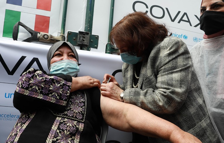 Palestinian health minister Mey Keyle gives a woman an injection. In the background are signs reading 'COVAX'.