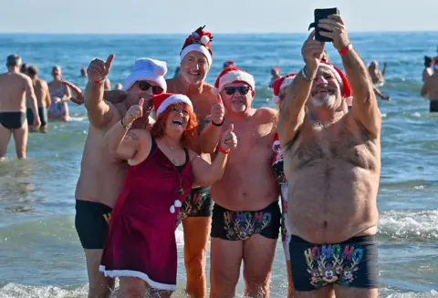 AFP via Getty Images A group of people in the sea wearing santa hats and swimwear, with their thumbs up taking a selfie