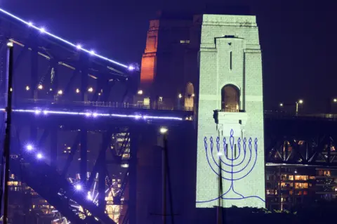 Getty Images An image of a menorah is projected onto the pylons of the Sydney Harbour Bridge during New Year’s Eve celebrations on December 31, 2025, in Sydney, Australia.
