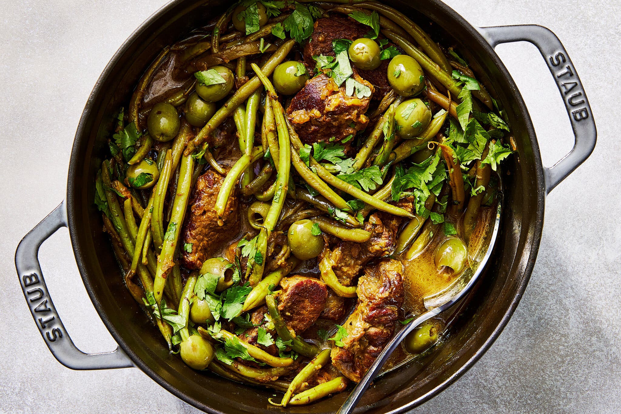 Large chunks of braised beef are nestled among bright green beans and whole green olives in a dark Staub Dutch oven, garnished with torn cilantro.