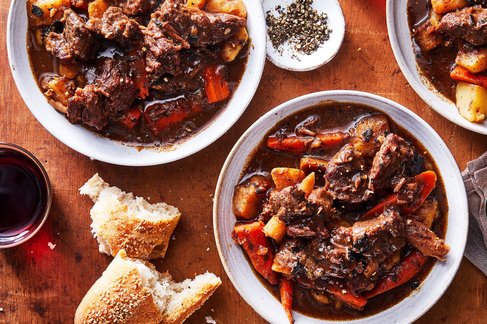 Bowls of hearty beef and carrot stew served on a wooden table with torn pieces of sesame baguette and red wine.