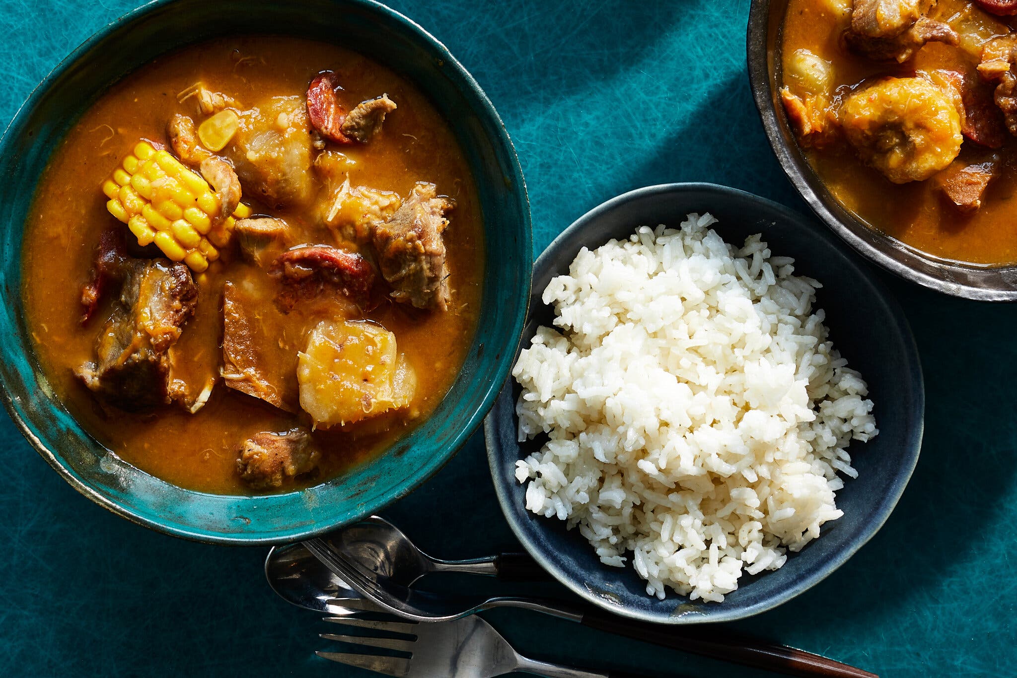 A teal bowl of golden-brown stew with corn and meat, served alongside a separate bowl of fluffy white rice.