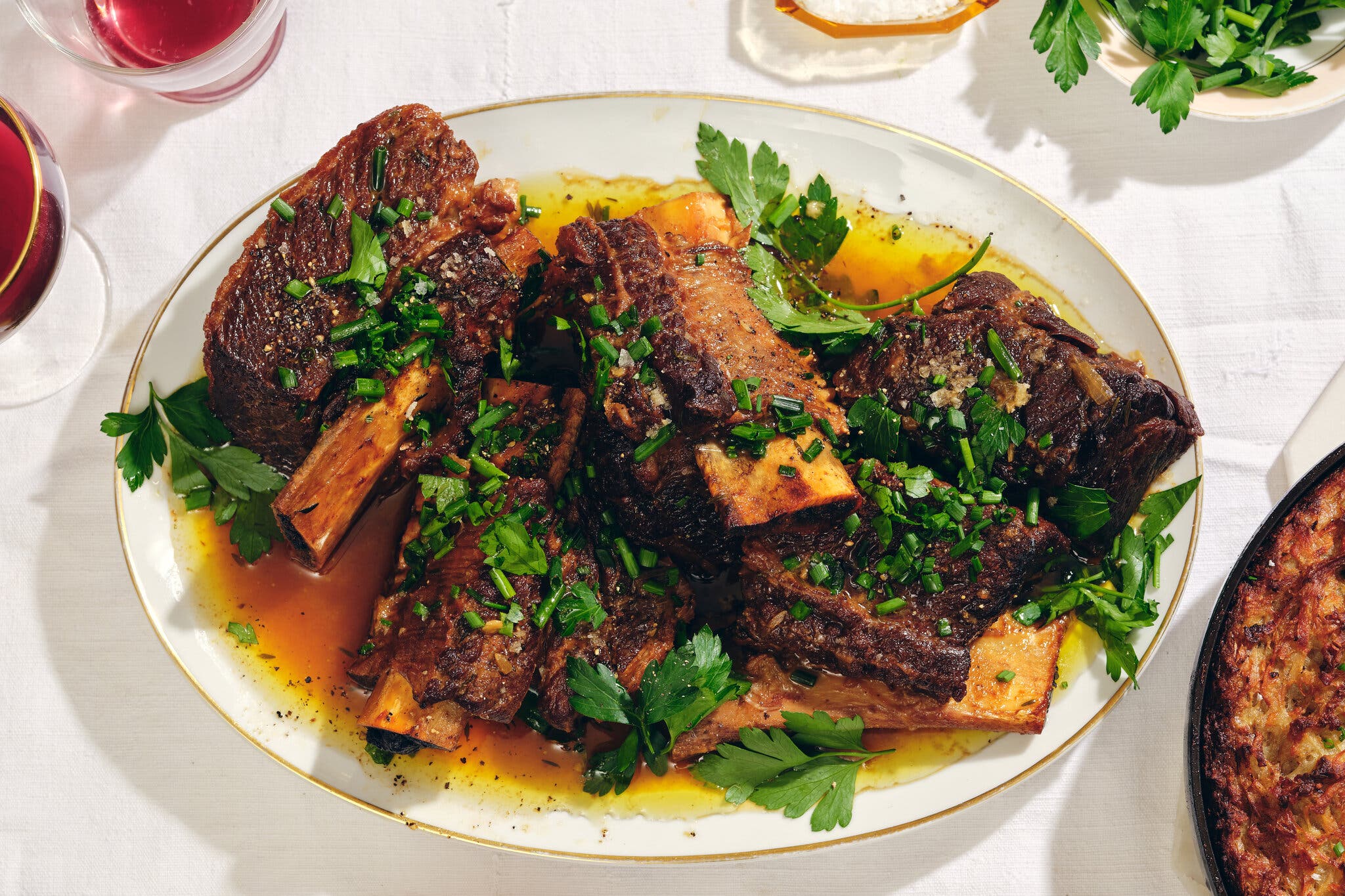 Braised beef short ribs on a white platter, garnished with fresh parsley and chives.