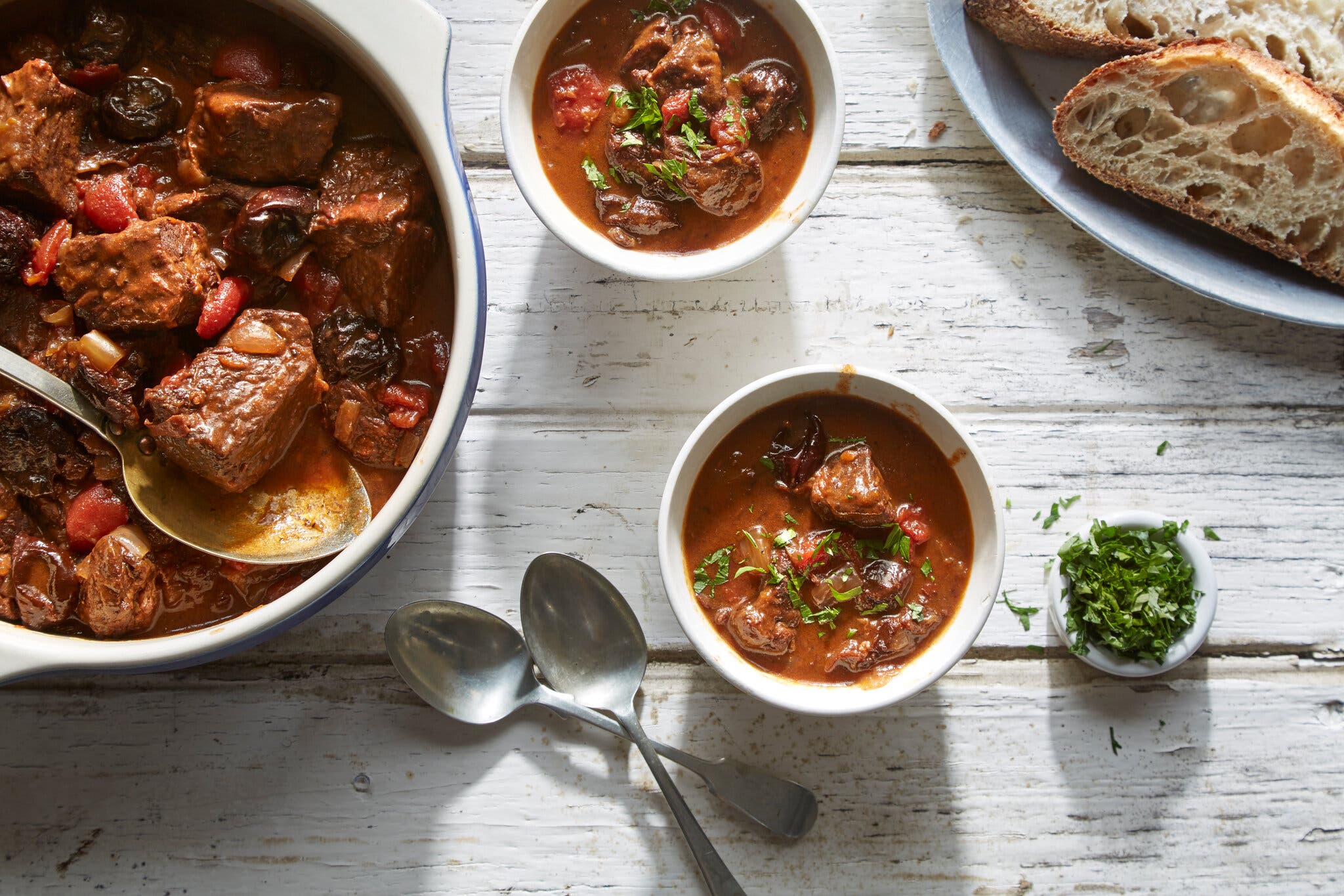 Bowls of beef and prune stew served on a rustic white wood table.