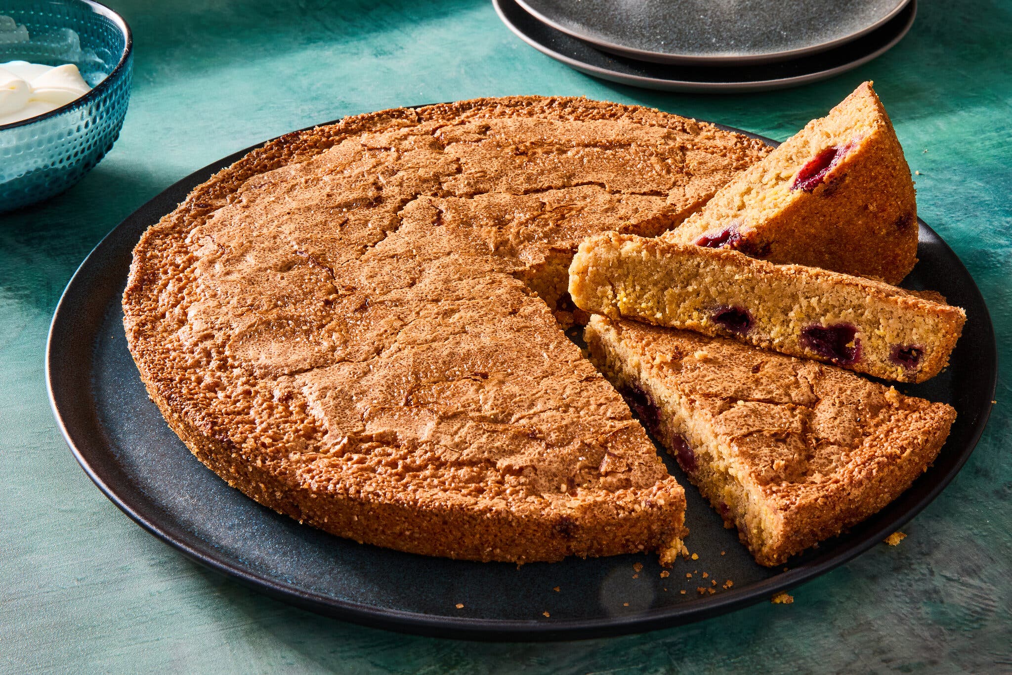 A round, golden-brown almond cake with dark cherries, sliced on a black platter.