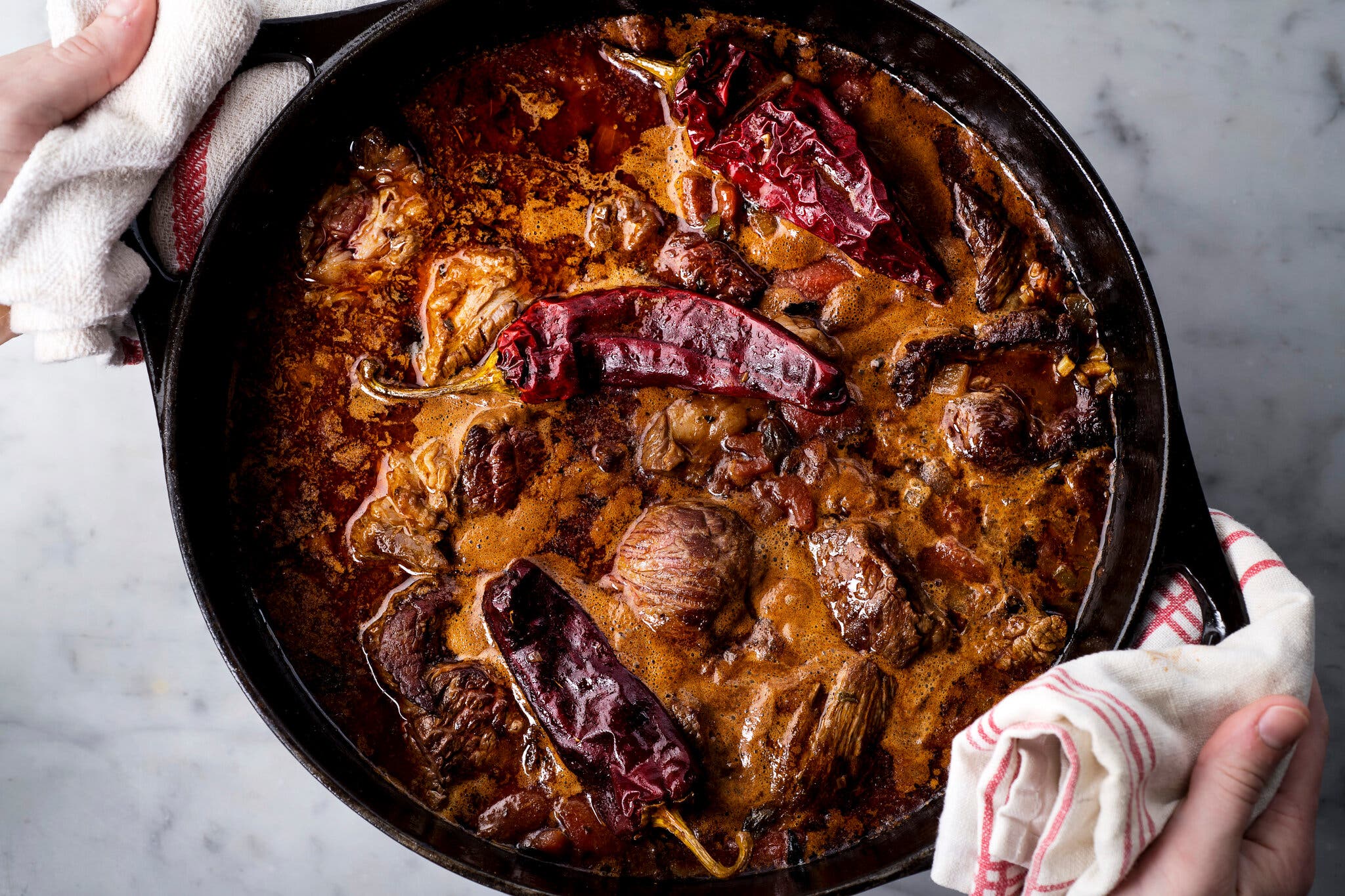 Chunks of beef and onions simmered in a thick, dark sauce, topped with several whole dried red chilies. A person’s hands hold the pot using white and red striped kitchen towels.