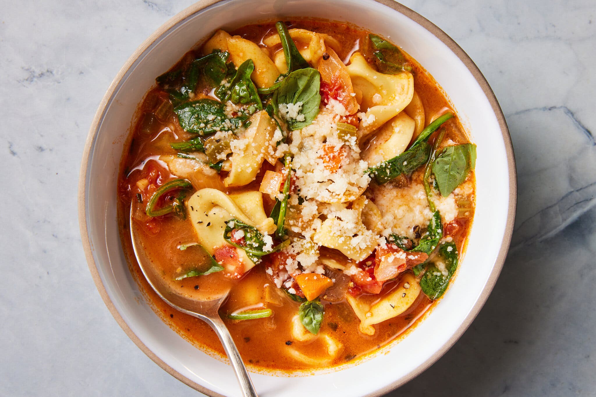 An overhead shot of a bowl of reddish soup with tortellini, chopped carrots, spinach and cheese.